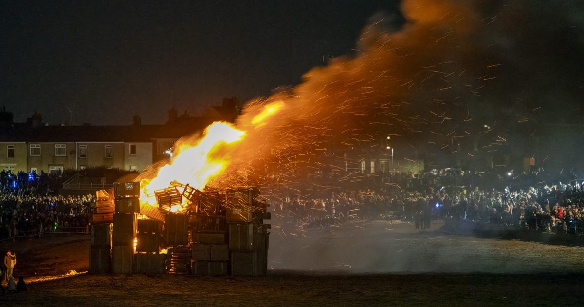 A large bonfire of stacked wooden pallets throws sparks into the night sky as a crowd watches from behind a barrier