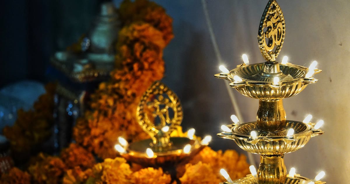 A tiered brass diya stand with lit lamps and an Om symbol in front of marigold garlands at a Lakshmi Puja altar