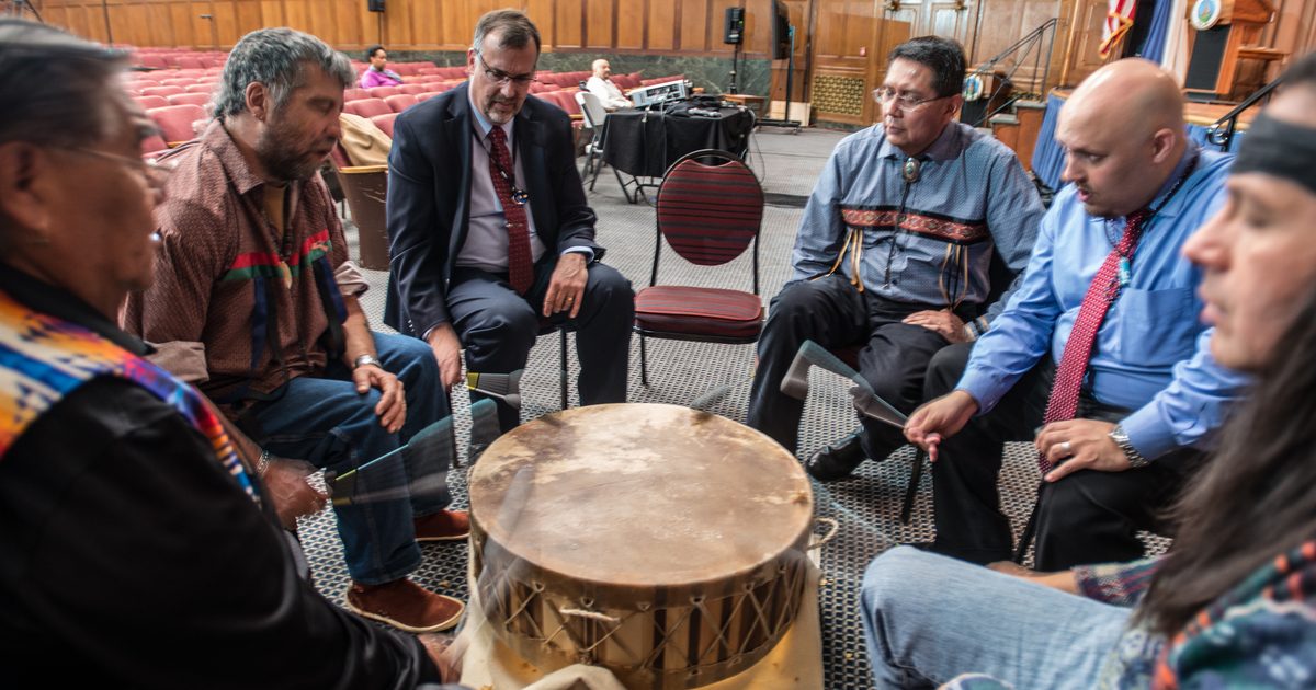 Six singers sit around a large powwow drum with drumsticks raised, practicing in an auditorium