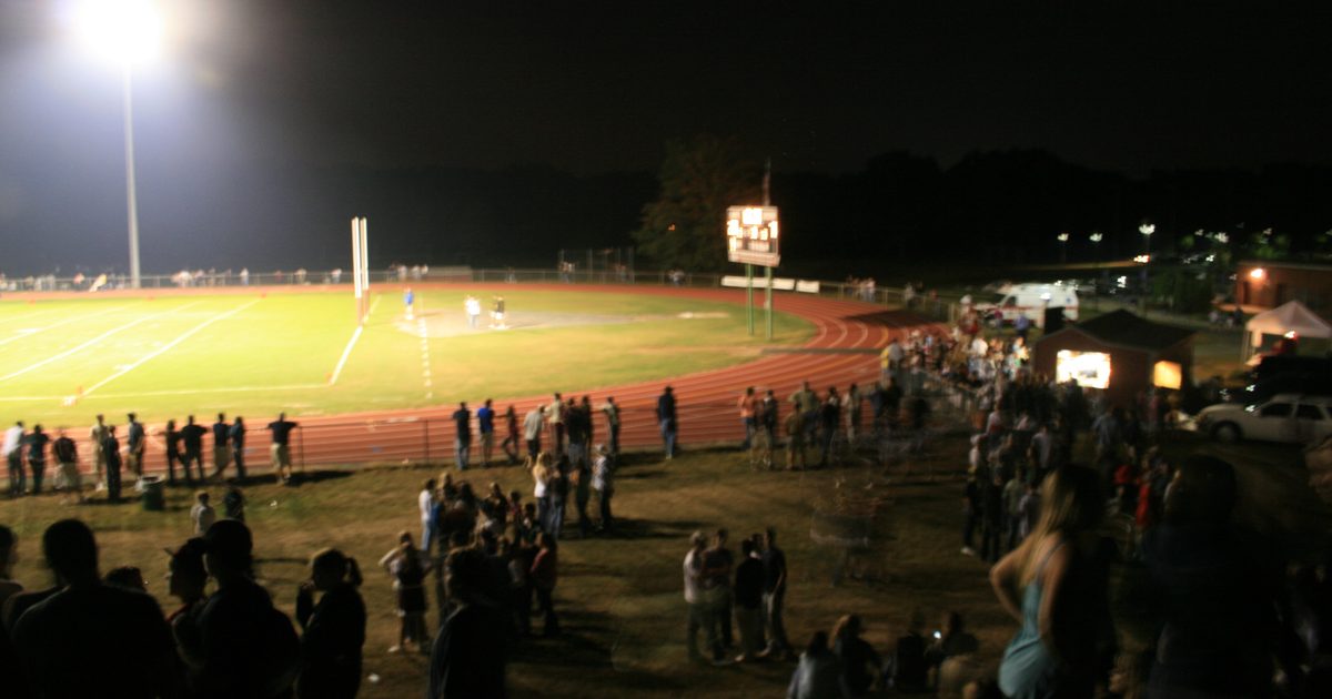 A high school football field under the lights at night, with families standing along the track and a crowd watching from the hill