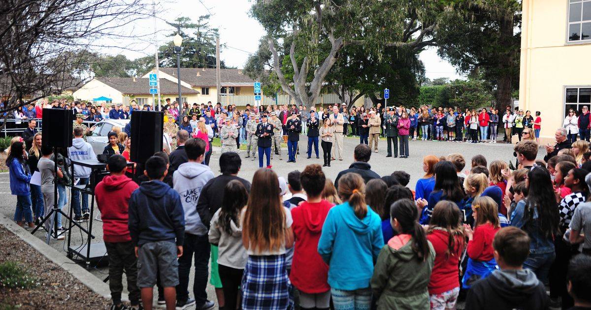 Students stand in a circle around veterans and service members at an outdoor school Veterans Day ceremony, with two speakers on stands
