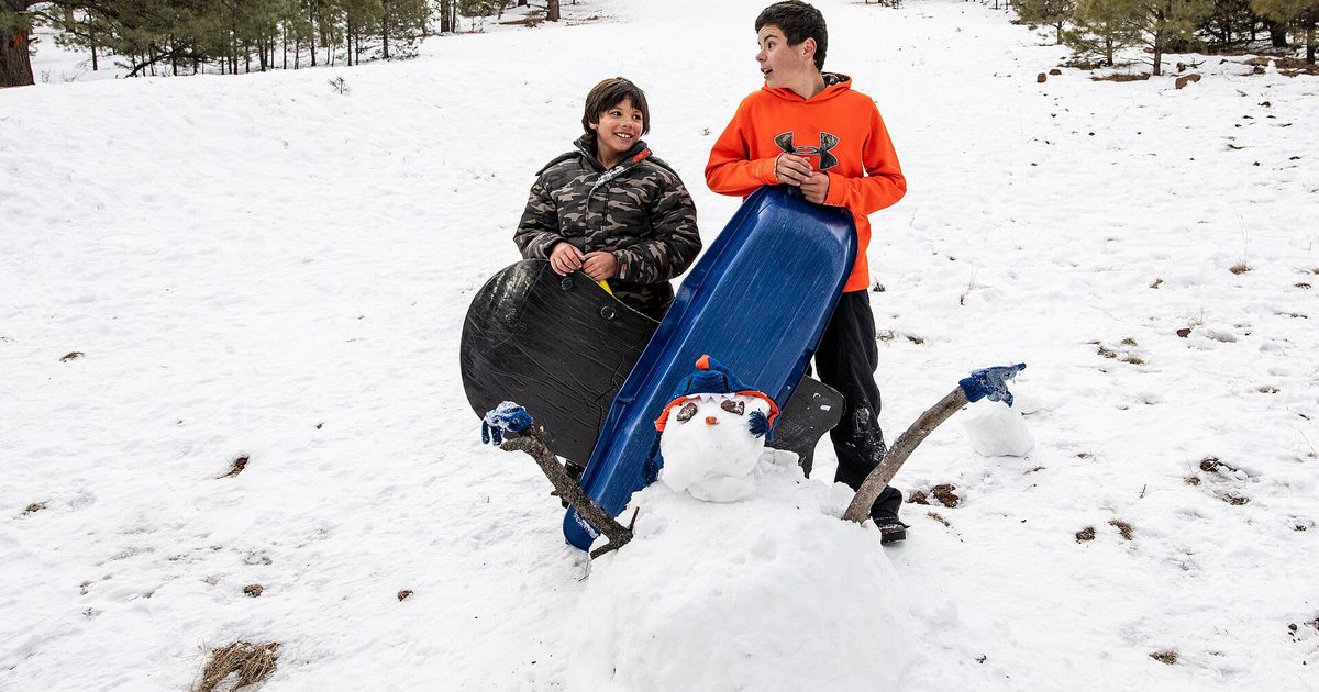Two boys holding a black saucer sled and a blue toboggan behind a small snowman on a snowy forest hill